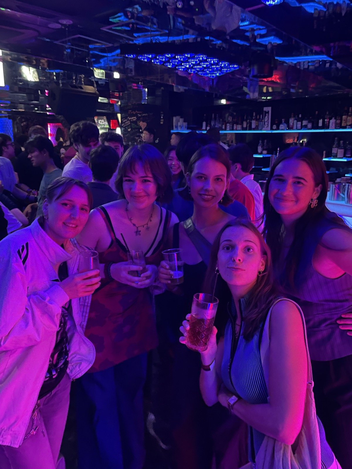 紫の照明のクラブでドリンクを手に笑う参加者たち — Friends holding drinks and smiling under purple club lights
