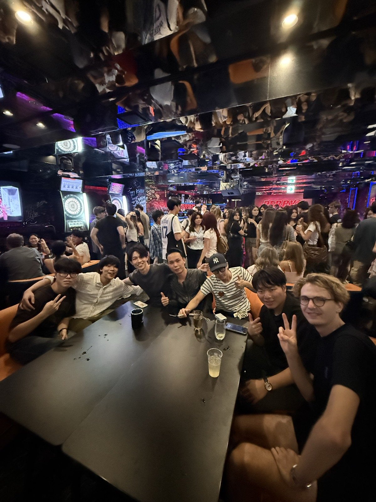 満員のバーでテーブルを囲みピースする学生たち — Students flashing peace signs around a table in a packed bar