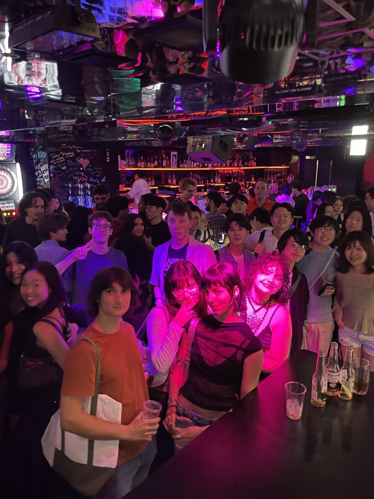 ピンクの照明の下、バーに集まって笑顔を向ける大勢の学生たち — A crowd of smiling students packed into a bar under pink lights